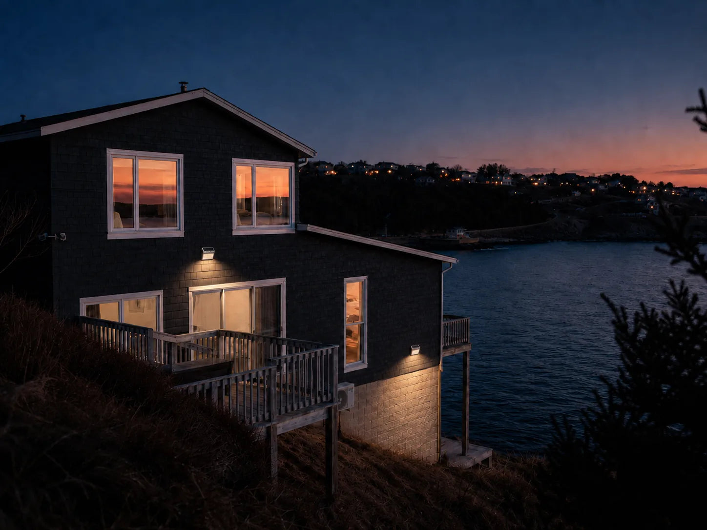 Two CampVale lights glowing on a dark shingle house above the bay at dusk
